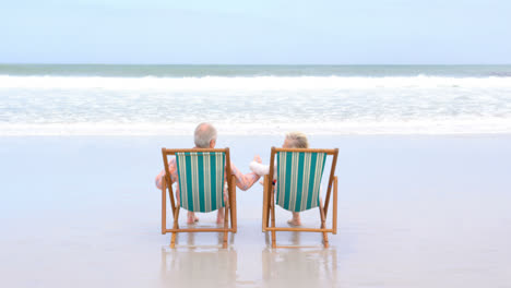 Rückansicht-Eines-Alten-Kaukasischen-Seniorenpaares,-Das-Auf-Einer-Sonnenliege-Sitzt-Und-Händchen-Hält-Am-Strand-4k