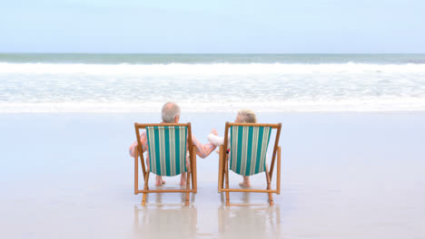 vista trasera de una vieja pareja de ancianos caucásicos sentados en una tumbona y tomados de la mano en la playa 4k