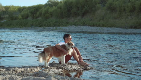 young man pets his dog on the river bank