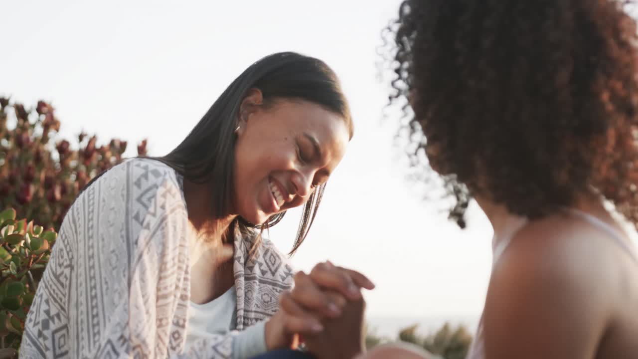 feliz pareja romántica de lesbianas biraciales sentadas tomadas de la mano en el jardín al atardecer, cámara lenta