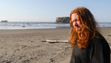 un hombre con el pelo largo y rojo mira hacia una playa en oregon contemplando tranquilamente