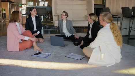 A-group-of-girls-in-business-suits-sit-on-a-gray-carpet-in-the-office-and-communicate-with-each-other-during-a-break-at-work-in-a-modern-office