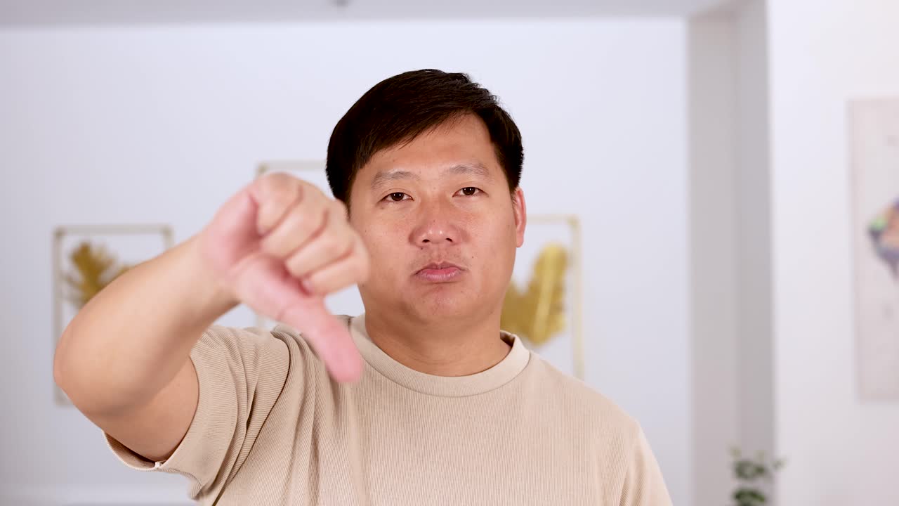 Middle-aged Asian man in beige shirt gives thumbs down gesture with serious expression in a bright, modern indoor setting. Static camera, soft lighting