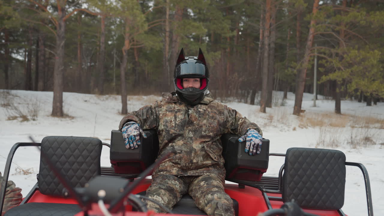 Pilota solitario seduto su un ATV rosso in una foresta di pini innevata, casco integrale con visiera oscurata e tuta mimetica, postura rilassata ma concentrata, guanti sulle manopole, motore al minimo, terreno ghiacciato e neve fresca in evidenza.