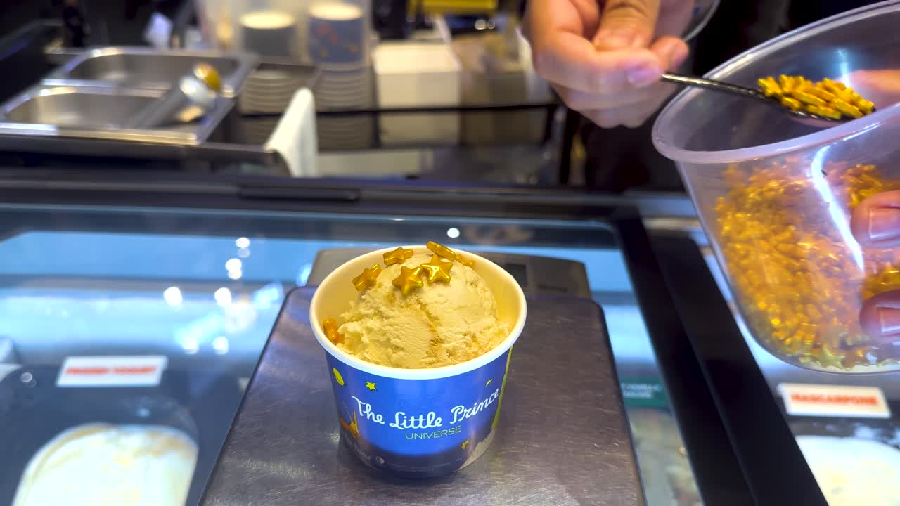 A hand adds crunchy toppings to ice cream in a Bangkok shop. Bright lighting highlights the detailed process