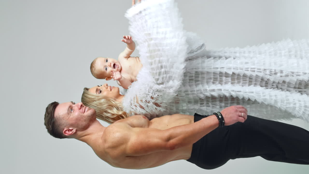 Athletic man with naked torso stands near blonde woman in white robe. Loving parents with their cute baby boy. Vertical screen. White backdrop.