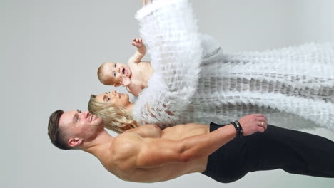 athletic man with naked torso stands near blonde woman in white robe. loving parents with their cute baby boy. vertical screen. white backdrop.