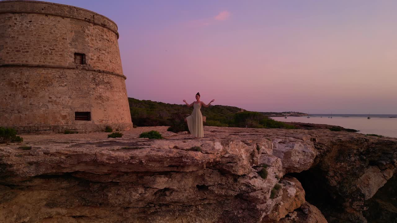 Woman performing yoga poses at sunset near Tower and Es Vedra Island in Ibiza, Spain. Unbelievable aerial view flight static tripod hovering drone
