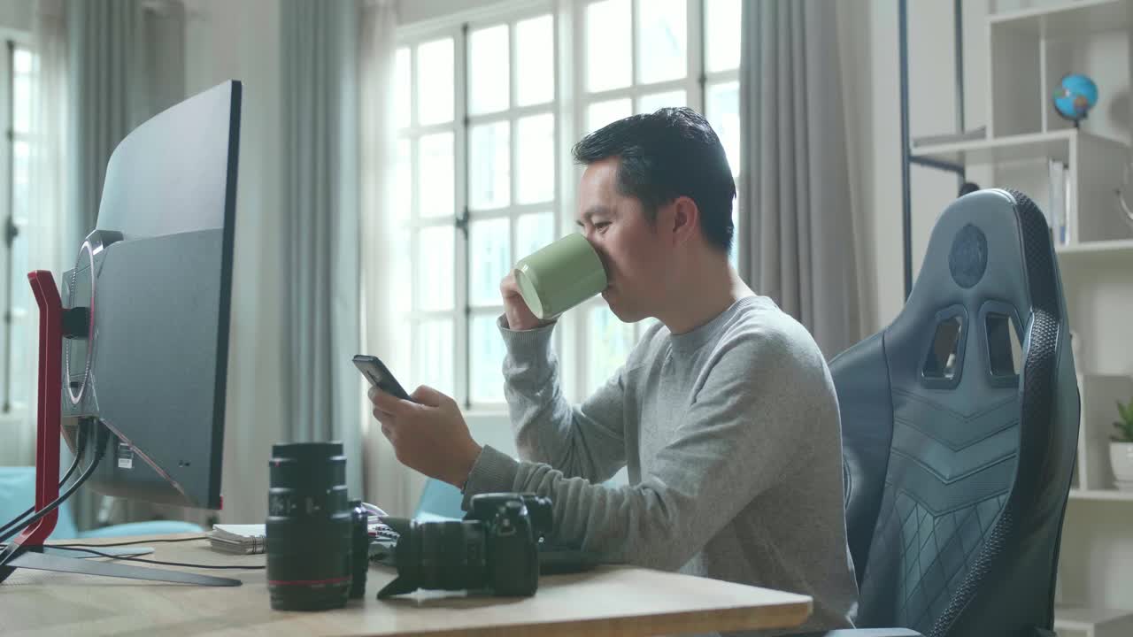 Asian Cameraman In Long Sleeved T-Shirt Using Mobile Phone And Drinking Coffee While Using Desktop Computer For Working At Home.