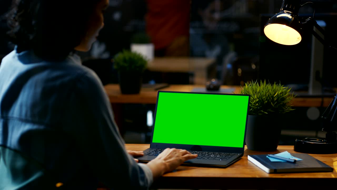 una trabajadora de oficina en su escritorio trabaja en una computadora portátil con una maqueta de pantalla verde. sobre las imágenes del hombro. se sienta en el escritorio de madera en la oficina creativa.