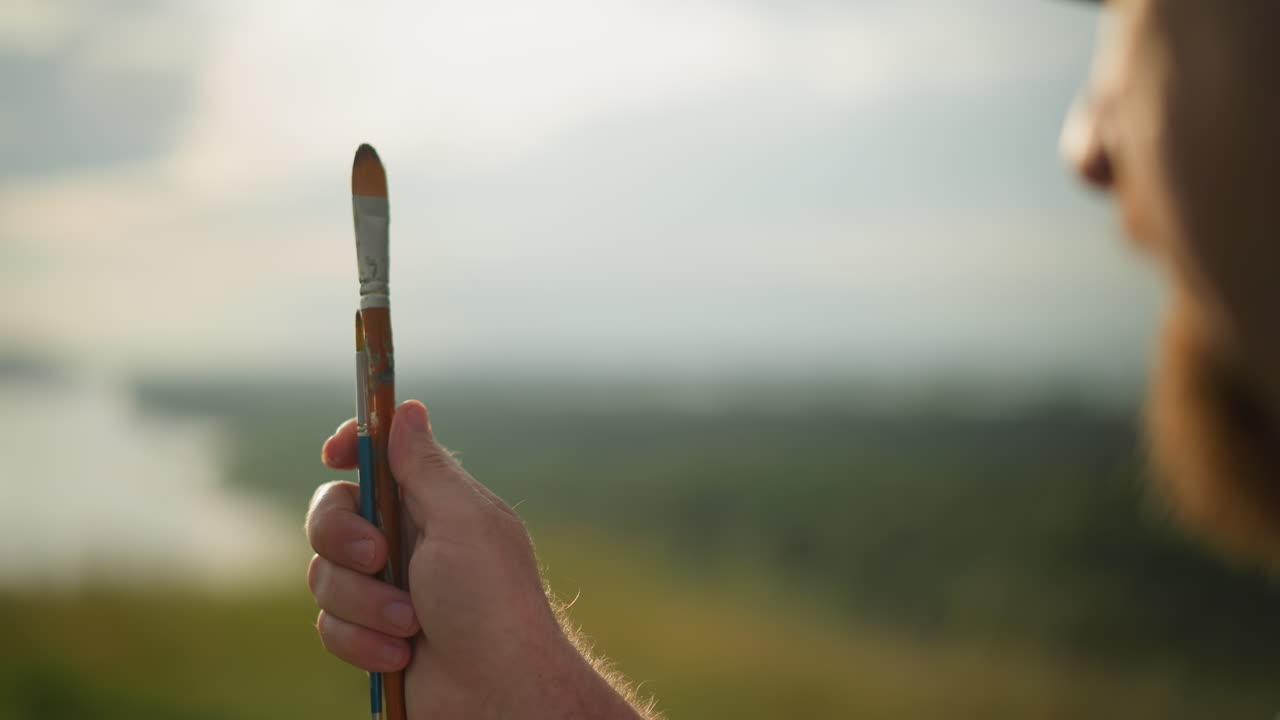 A close-up of a person's hand holding two paintbrushes one small and one very small while turning them slowly. The face is partially visible against a blurred, sunny background