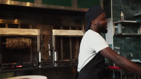 a black person dances at work in a doner market against the backdrop of a barbecue. man in black apron and white t-shirt having fun at work