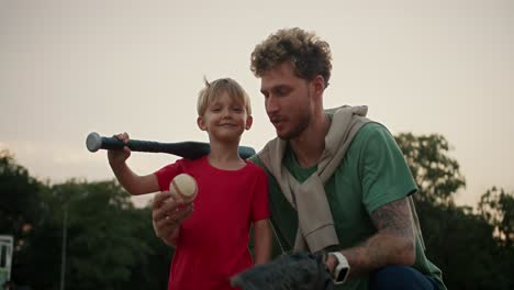 Happy-little-blond-boy-in-a-red-T-shirt-holds-a-black-baseball-bat-in-his-hands-and-throws-a-baseball-into-his-dad's-baseball-glove-in-a-Green-T-shirt.-Dad-and-son-playing-baseball-in-the-park