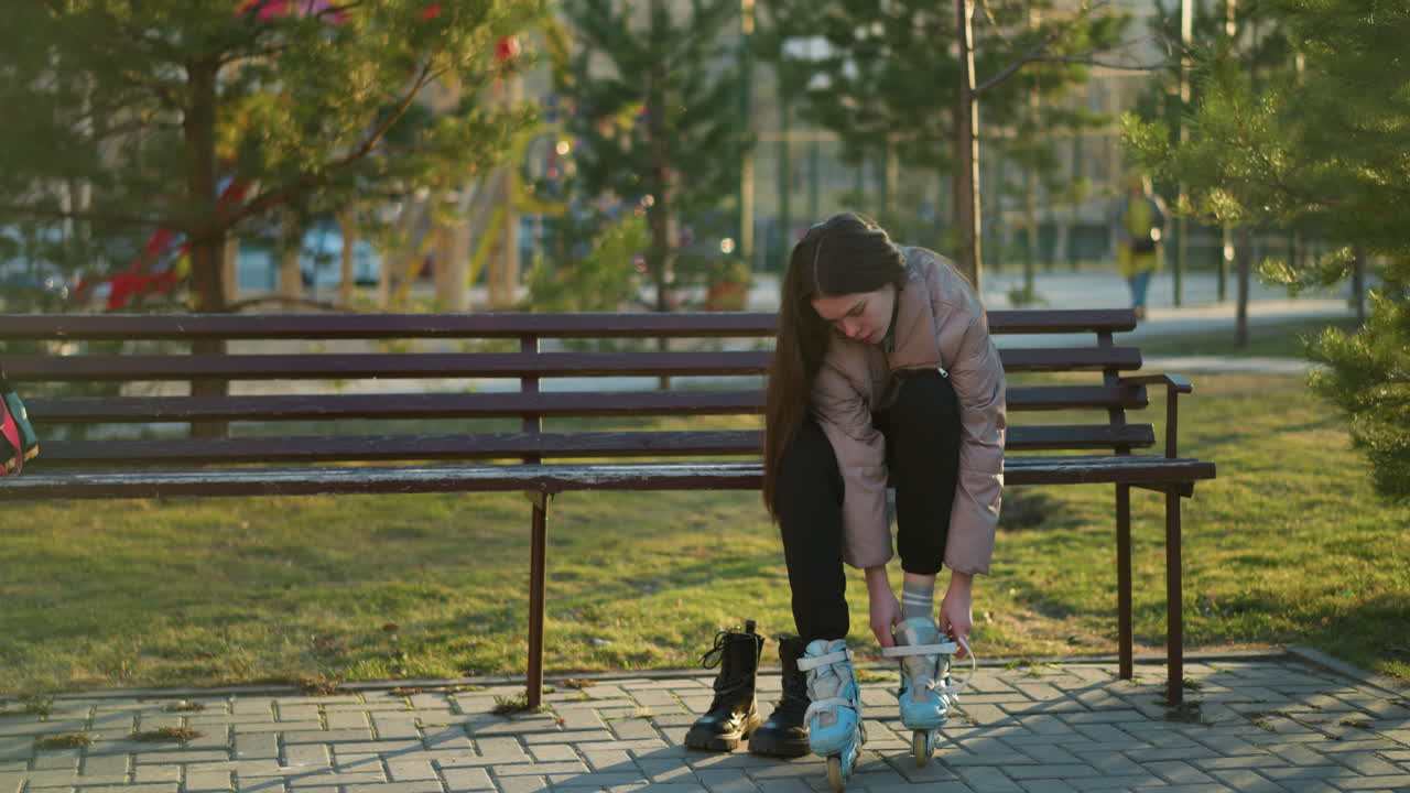 A girl in a peach jacket and black trousers sits on a bench in a park, picking up a rollerblade from the bench to put it on. moment of preparation for a skating session in an outdoor park setting