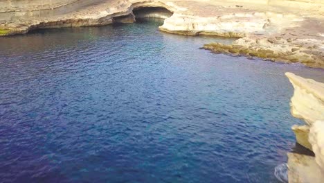 Ein-Flug-über-Den-Malerischen-St.-Peters-Pool-Auf-Malta-Mit-Blick-Auf-Die-Alten-Salzpfannen,-Die-In-Die-Goldenen-Sandsteinfelsen-Gehauen-Sind,-Und-Das-Kristallklare-Blaue-Wasser-Des-Mittelmeers