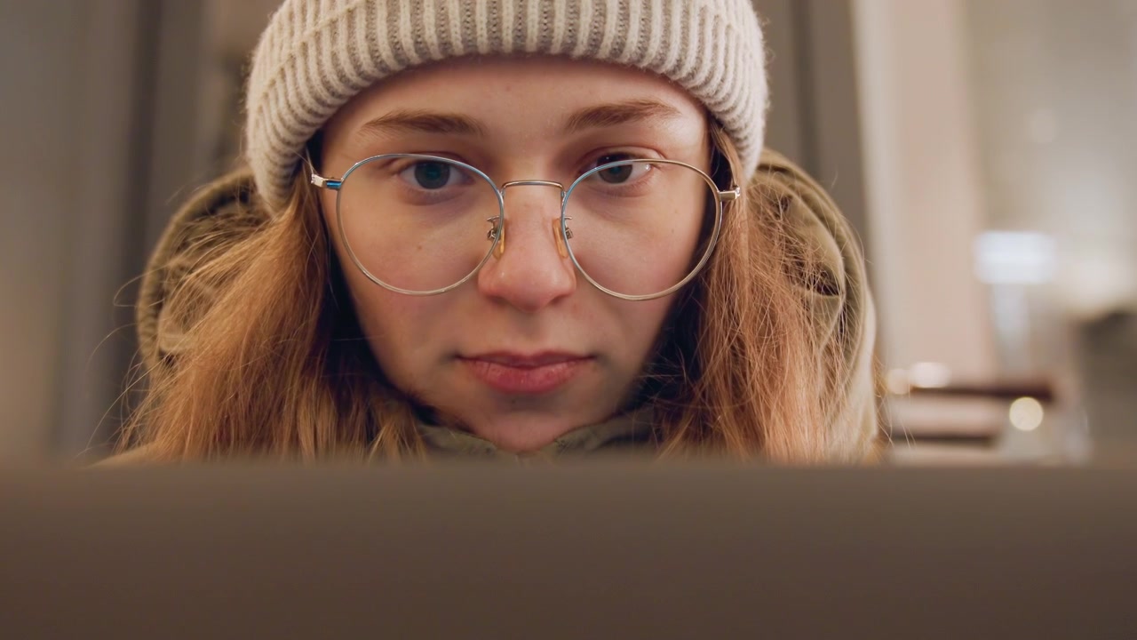 Junger Student lernt am Laptop unter warmem Licht, konzentriert auf Aufgabe, trägt Mütze und Brille, Winterjacke, Nahaufnahme von Bildschirm und tippenden Händen, Umgebungs-Bokeh und Kaffeehintergrund