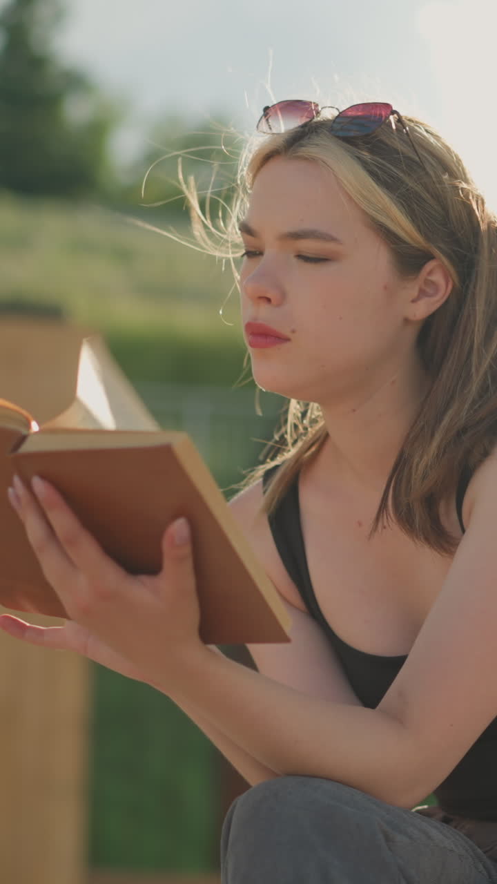 mujer sentada, leyendo un libro mientras el viento sopla su cabello y páginas, su mano descansa en el libro con una bolsa negra cerca, y el fondo presenta una colina de hierba borrosa en la luz del sol
