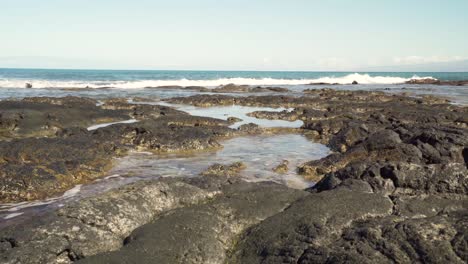 Pequeñas-Olas-Que-Vienen-Hacia-La-Cámara-Cubriendo-Una-Formación-De-Roca-De-Lava