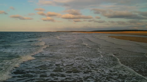 Low-Aerial-Drone-Shot-at-Golden-Hour-over-Small-Waves-Breaking-on-Empty-Sandy-Beach-Coastline-with-Golden-Sand-UK