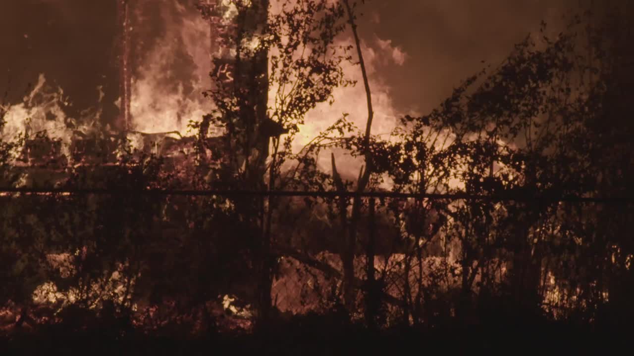 Flames tear through a structure behind silhouetted trees and fence, as the night sky glows from the heat and smoke
