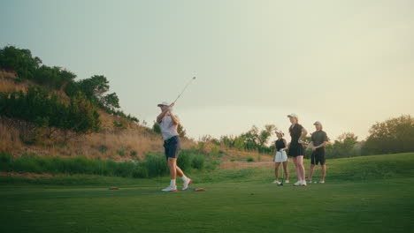 Golf foursome gathers on the tee box in golden hour light as a male golfer hits an iron shot. After the swing, the group celebrates with a cheerful high five in a relaxed, scenic moment.