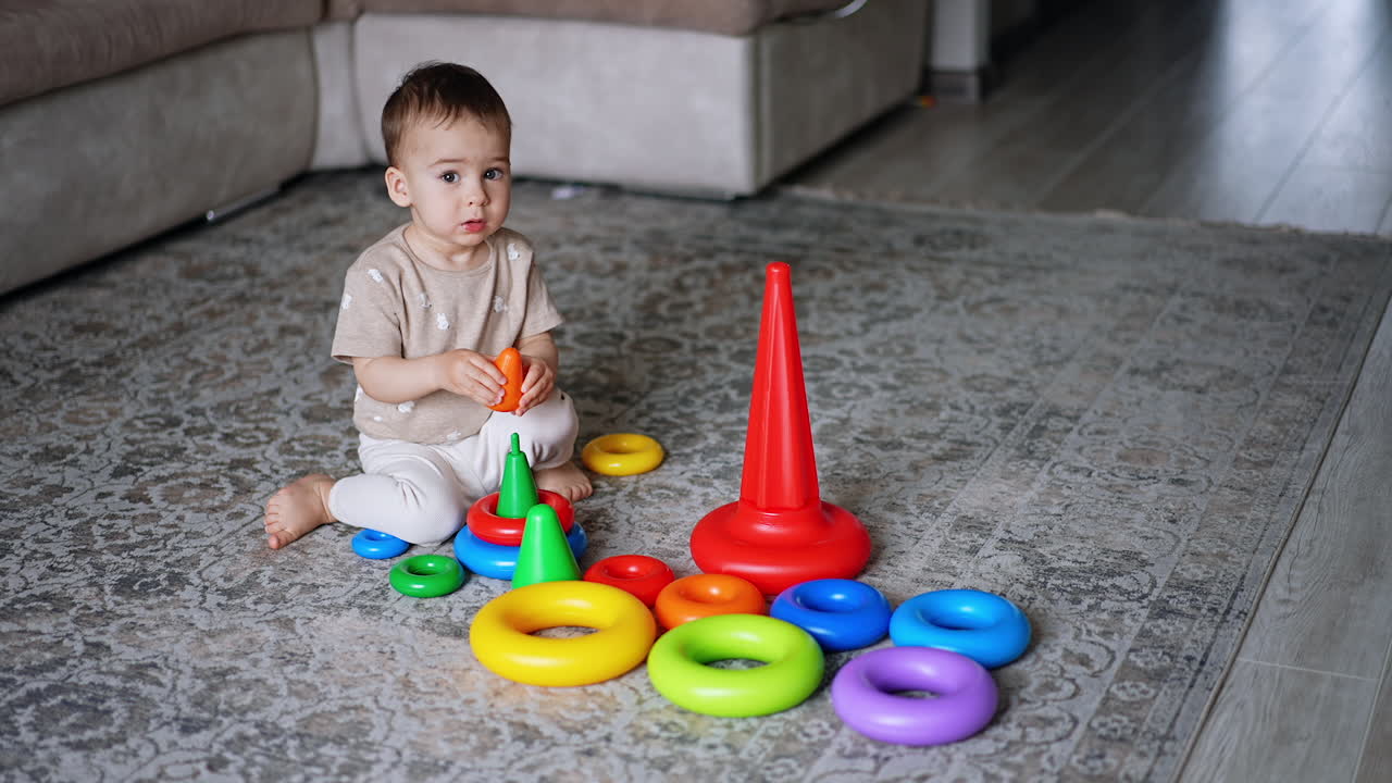 Little barefoot boy sits on the carpet surrounded by toys. Caucasian toddler learning colors assembling the pyramid.