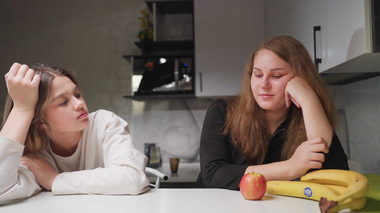 Medium view of girlfriends seated calmly and thoughtfully at kitchen table with apple and banana in front, cabinets behind them, relaxed indoor atmosphere showing quiet moment of reflection