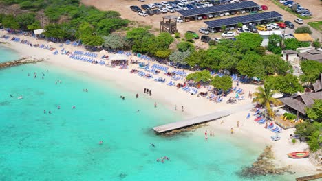 Drohne-Umkreist-Den-Pier-In-Playa-Porto-Mari,-Während-Touristen-Am-Weißen-Sandstrand-Schwimmen-Und-Spazieren-Gehen