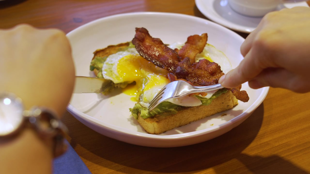 Persona che taglia un toast salato con avocado, uovo fritto e pancetta croccante su un piatto bianco a un tavolo di legno di un caffè.
