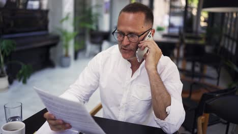 an adult man in a shirt walks through a restaurant with a paper and speaks on the phone