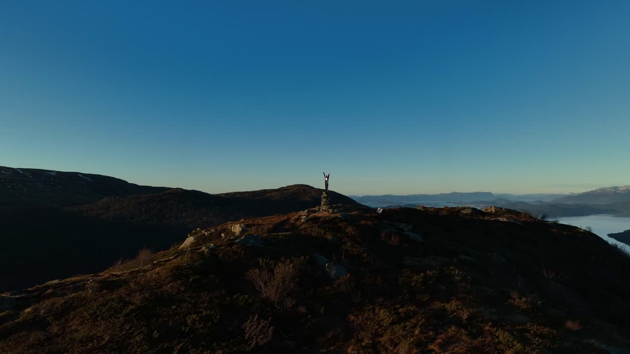 Uomo sulla cima di una montagna che alza le braccia in segno di vittoria. Drone che orbita catturando silhouette e viste illuminate dal sole
