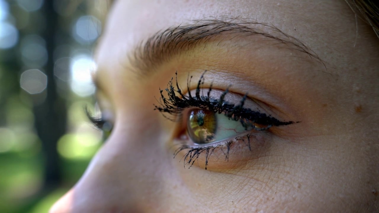 Blinking woman's eye reacting to bright glare at park, showing winged eyeliner and clumped mascara