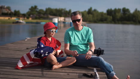 father and son take a selfie with the american flag on a lake pier