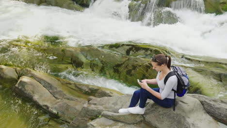 una persona usa un teléfono inteligente en una caminata sentada junto a un hermoso río de montaña