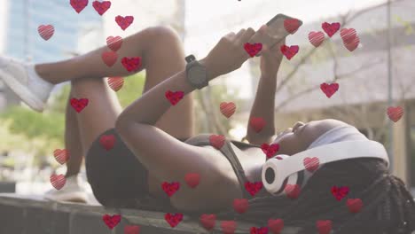 animation of heart icons over african american woman using smartphone outdoors