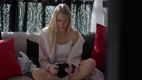 beautiful blonde white caucasian girl women sitting on couch in front of windows in pink bath robe resting relaxing at home playing video games with controller in hand smiling looking straight at tv