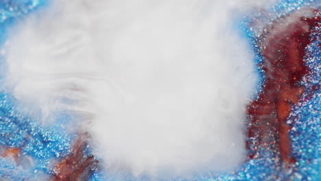 macro view of brown glitter ink blending with swirling white smoke in water. the visual creates soft, cloudy shapes framed by shimmering blue and orange accents