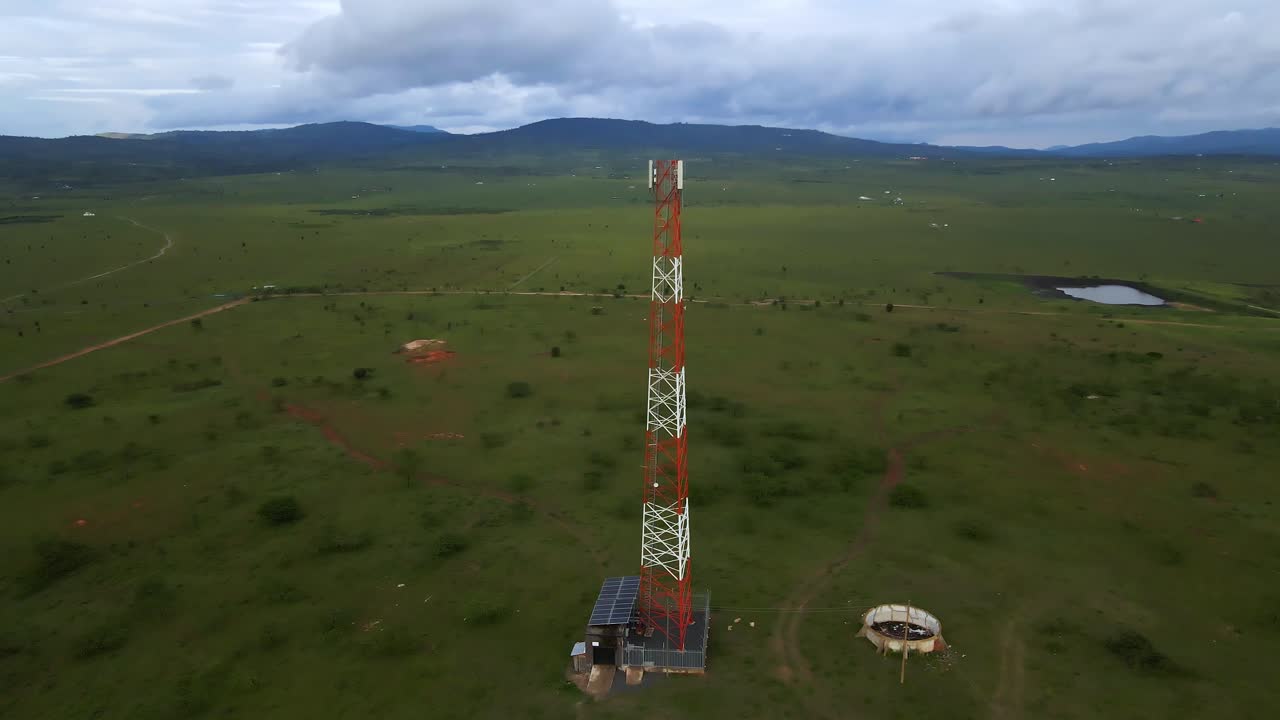 An aerial view of a cell tower standing tall in a vast, undulating green field, surrounded by a few trees and hills. The scene suggests connectivity and rural development