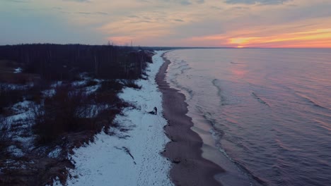 Statische-Luftaufnahme-Eines-Einsamen-Mannes,-Der-Am-Schneebedeckten-Strand-Von-Daugavgriva-Steht,-Während-Die-Wellen-Bei-Sonnenuntergang-Sanft-Am-Ufer-Brechen