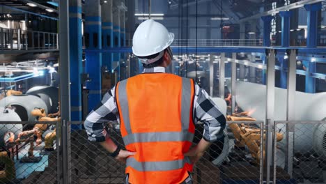 back view of asian male engineer with safety helmet in factory manufacture of wind turbines. standing with arms akimbo looking around, checking