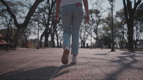 mujer camina tranquilamente por el parque, en el camino bordeado de árboles, con el sol brillando, proyectando sombras mientras pasea