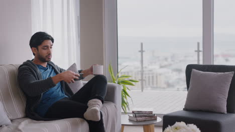 happy young man using smartphone browsing online reading social media messages enjoying comfortable lifestyle drinking coffee relaxing on couch at home