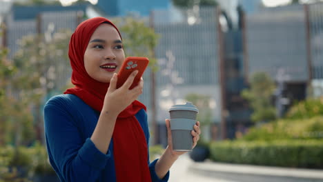 young muslim woman sending voice message on smartphone outdoors with coffee cup