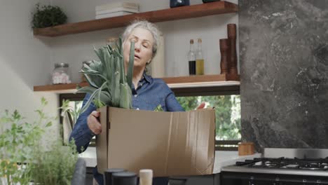 Happy-senior-caucasian-woman-holding-box-of-fresh-fruit-and-vegetables-in-kitchen,-slow-motion