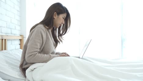 아름다운 미소 짓는 젊은 아시아 여성은 집에서 침실에서 침대에 앉아 노트북에서 일합니다.