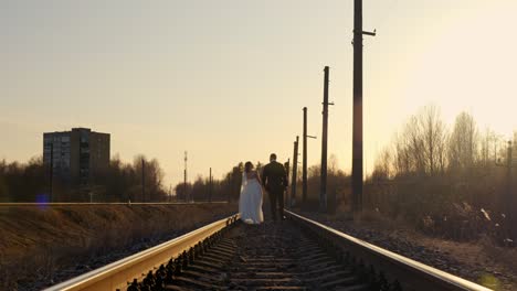 a wedding couple walks hand-in-hand on train tracks, bathed in the warm, golden sunset light.