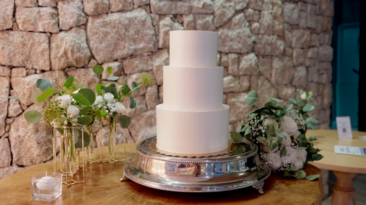 Bánh cưới ba tầng màu trắng trên giá đỡ bạc bên cạnh đồ trang trí hoa màu xanh lá cây dựa vào tường đá.