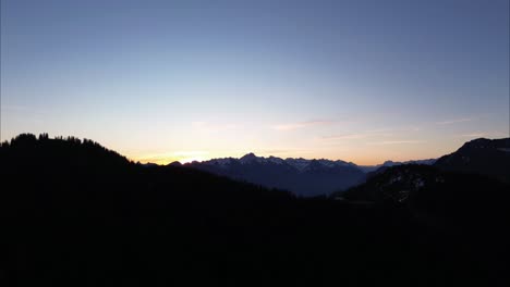 orange yellowish sunrise behind snow covered mountain range, with mountains and forest in foreground