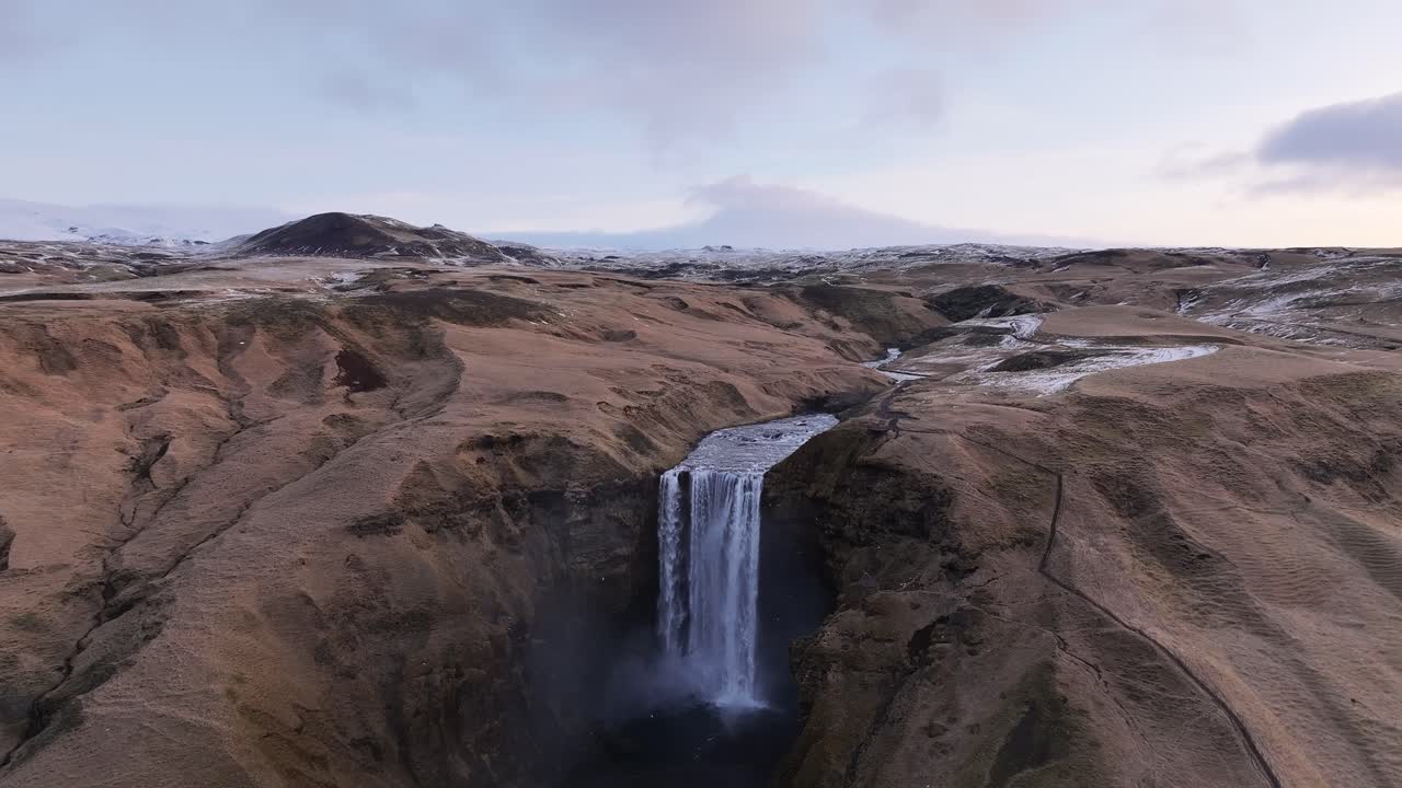 Aerial drone view of Skógafoss waterfall, Iceland, flowing from Skógá river near Eyjafjallajökull, surrounded by rugged terrain, mist spray and light snow in early winter.