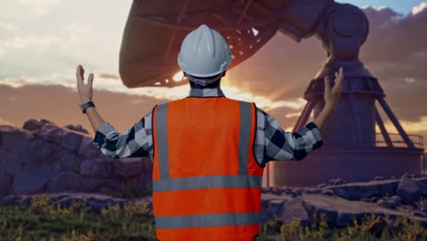 back view of a male engineer with safety helmet spreading arms while standing with large satellite dish